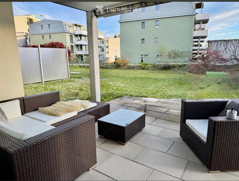 A modern outdoor patio with wicker furniture, including a sofa, chairs, and a small table, set on a tiled surface with a grassy area in the background.