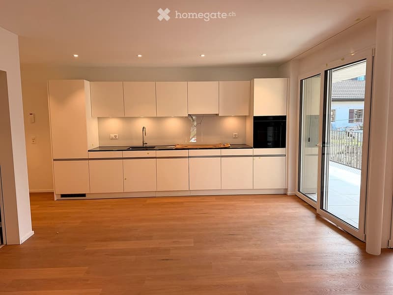 Modern kitchen with light-colored cabinets, a black countertop, and a wooden floor. Features a sink, faucet, and an oven, with a large sliding glass door leading outside.