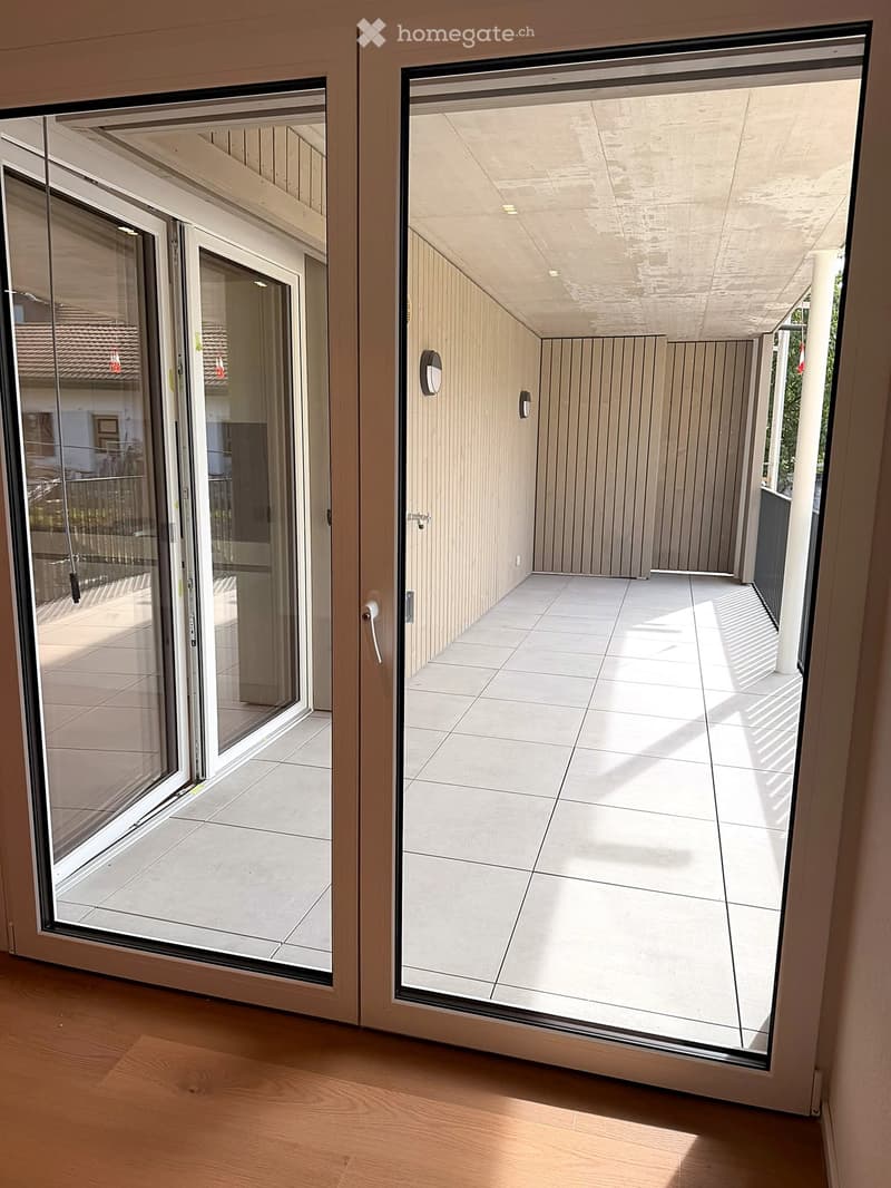 Open sliding glass doors lead to a modern balcony with tiled flooring, wooden panel walls, and a covered ceiling.