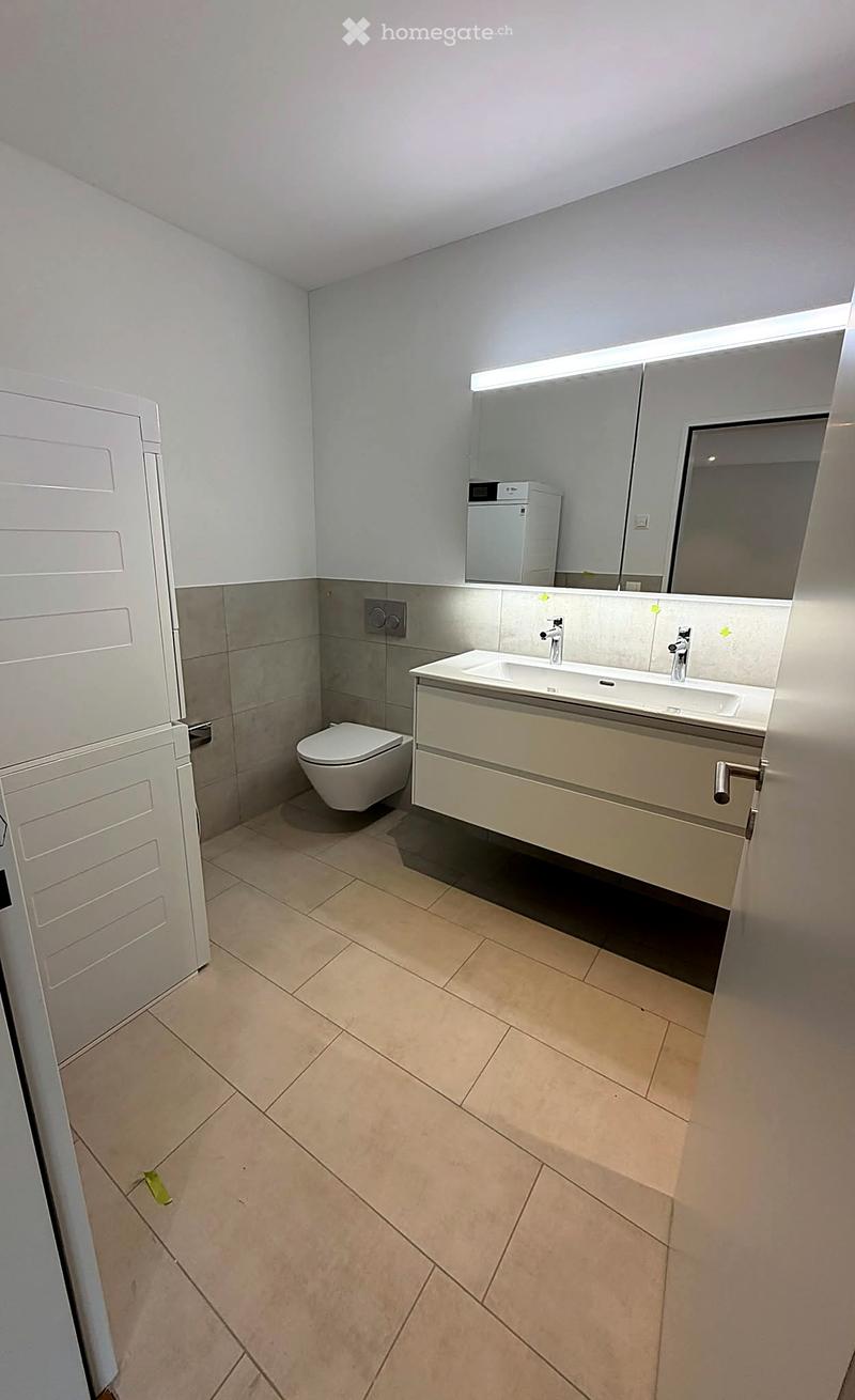 Modern bathroom with beige tiles, a double sink with chrome faucets, a large mirror with built-in lighting, and a toilet. The walls are painted white with a grey accent strip.