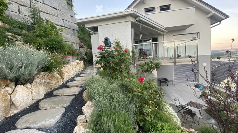 a white house with red brick wall, stone pathway, flowers and plants, balcony, table and chairs