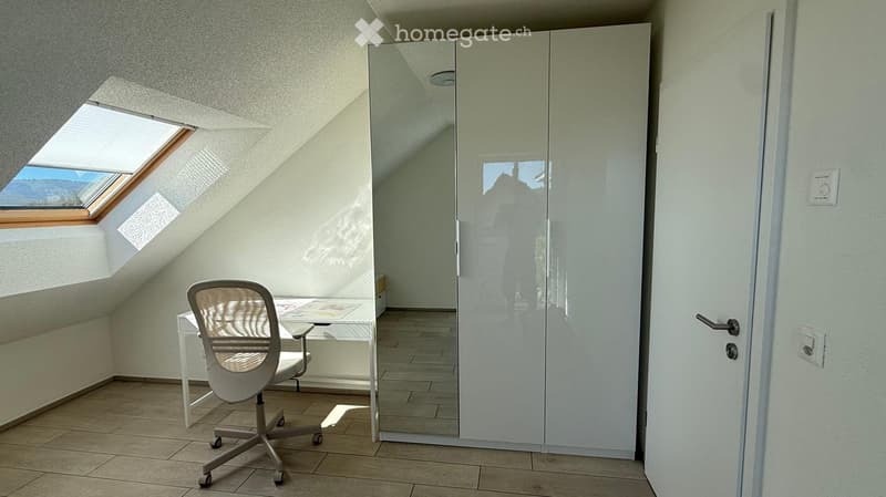 Modern, minimalist room with a slanted ceiling, large skylight window, white desk, ergonomic chair, and a large mirrored wardrobe.