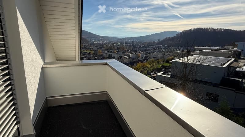 Balcony with white railings, overlooking a cityscape with buildings and distant mountains.