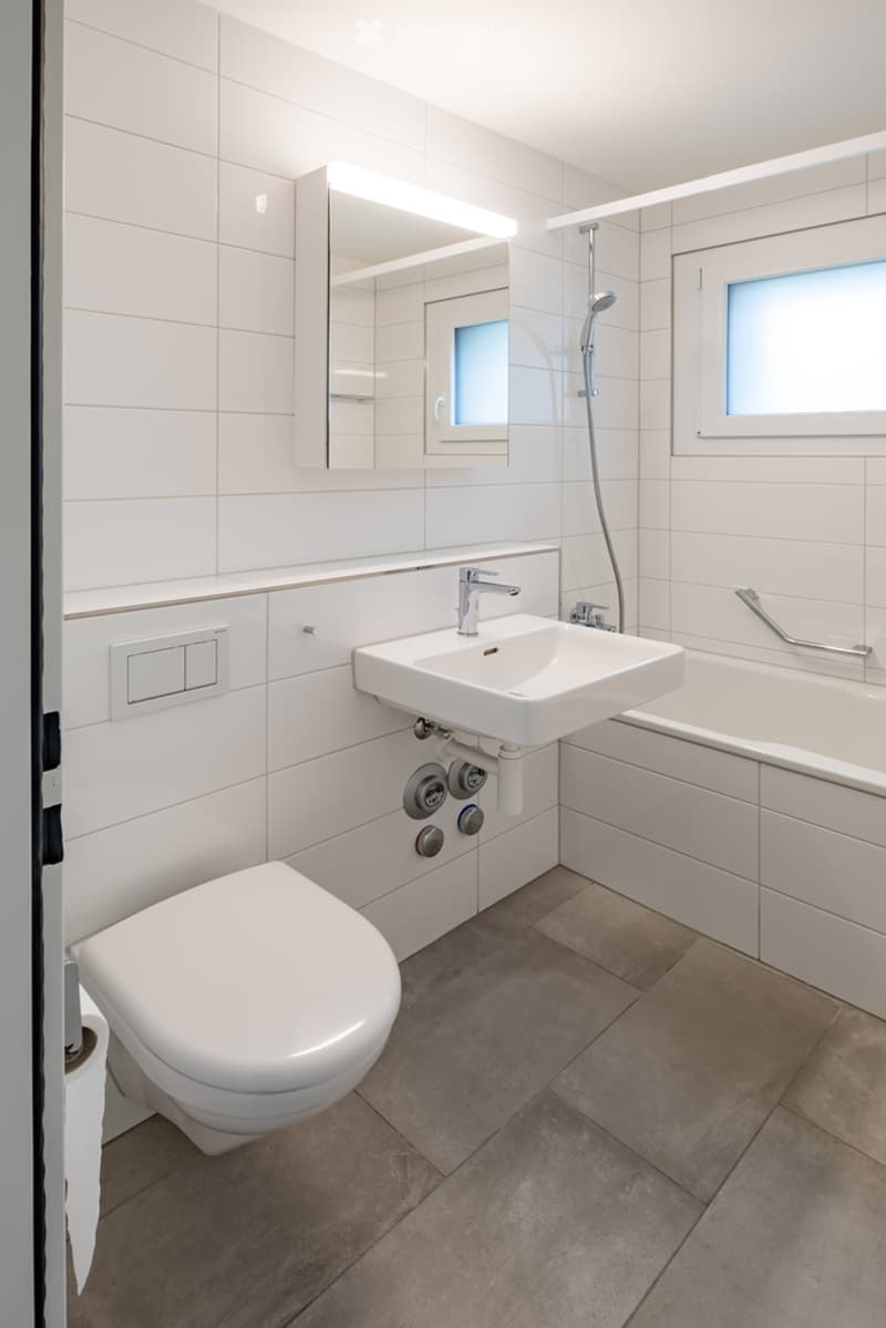 Modern bathroom with white tiles, a toilet, a sink, a bathtub, and a mirror with built-in lighting.