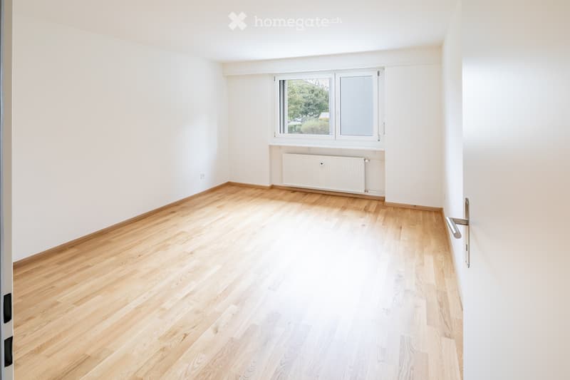 Empty room with light wooden flooring, white walls, and a window with a view of greenery outside. A radiator is visible below the window.