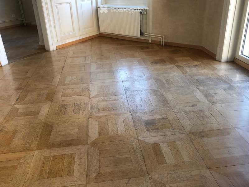 Empty room with wooden herringbone flooring, white walls, and a radiator visible on one wall.