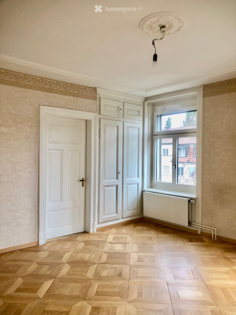 Empty room with wooden parquet flooring, white walls, a large window with a radiator below, and a white door.