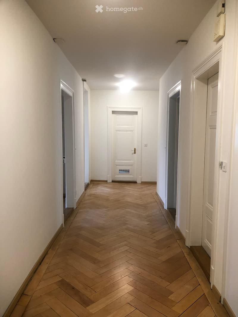 A narrow hallway with herringbone-patterned wooden flooring, white walls, and several doors leading to other rooms.
