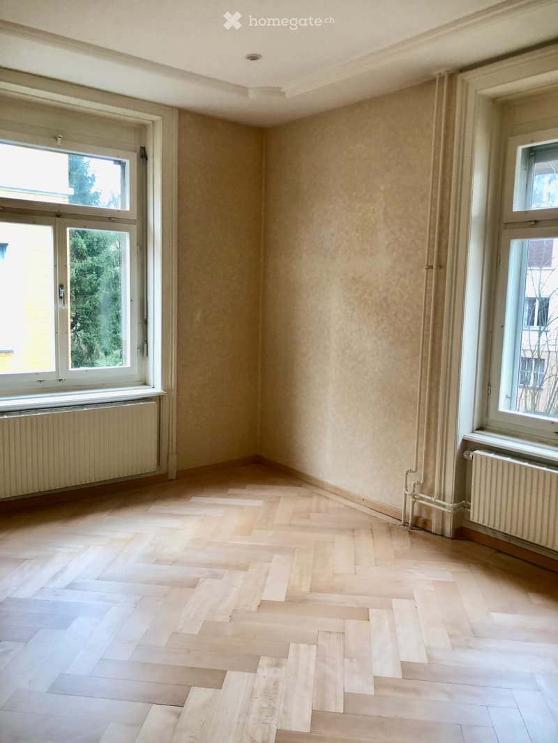 Empty room with light wooden herringbone flooring, beige walls, large windows with white frames, and radiators beneath the windows.