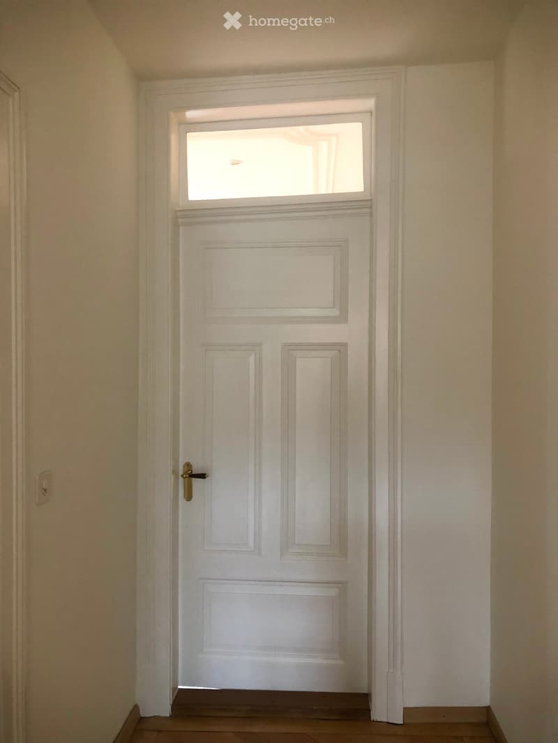 A white door with a transom window above it, set in a hallway with light-colored walls and wooden flooring.