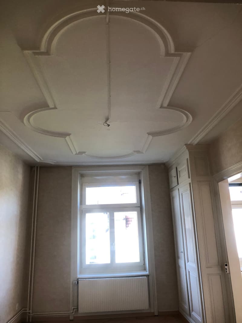 Empty room with beige walls, a large window with white frame, radiator below the window, and a white door on the right side. The ceiling has decorative molding and a central light fixture.
