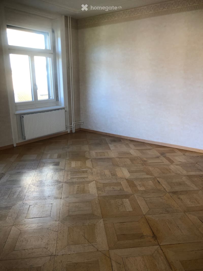 Empty room with wooden parquet flooring, white walls, and a window with a radiator below it.