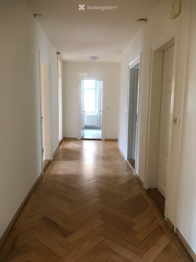 Empty hallway with wooden herringbone flooring, white walls, and several doors leading to other rooms.