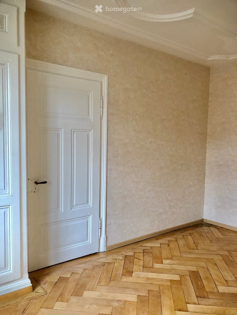 Empty hallway with wooden herringbone flooring and white walls, featuring a closed white door on the left side.