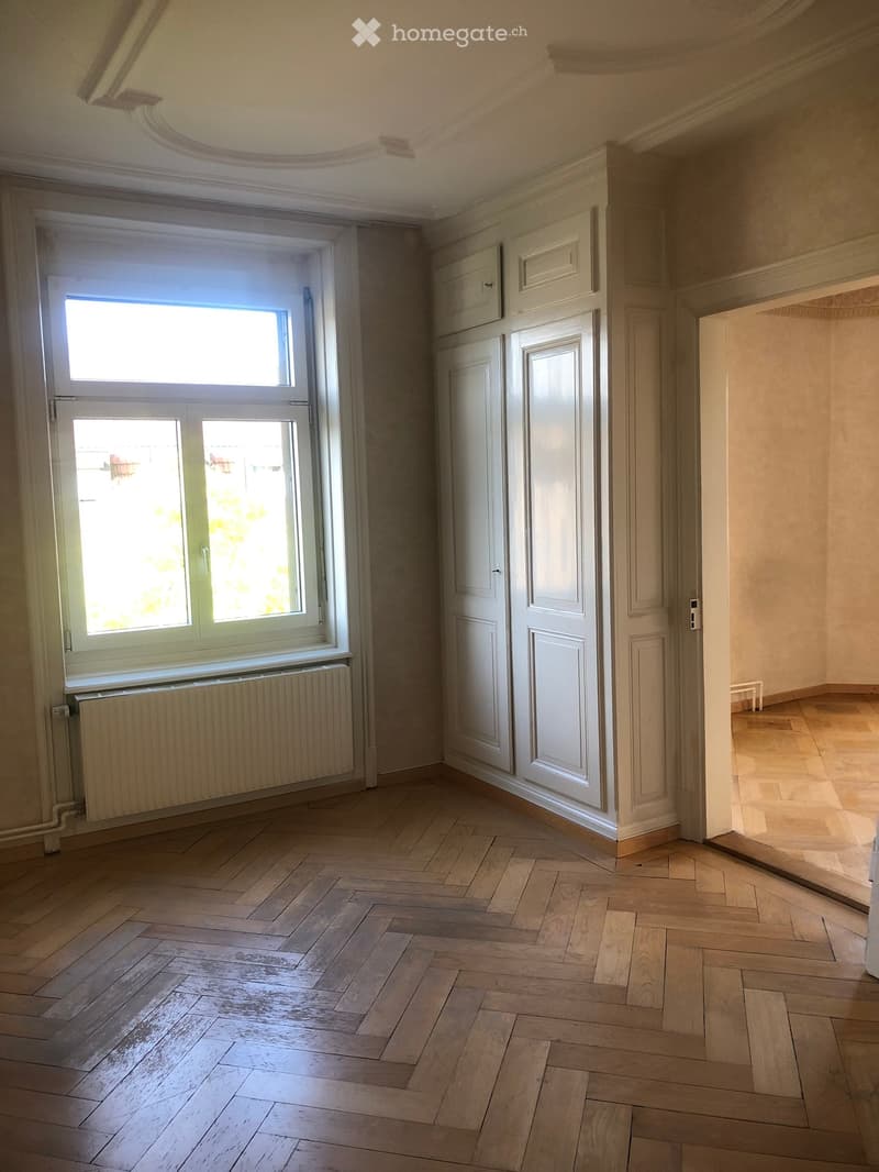 Empty room with wooden herringbone flooring, white walls, large window with radiator below, and several closed white doors.