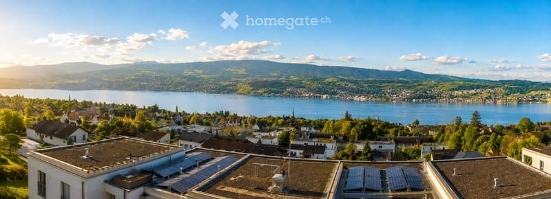 Panoramic view of a town with houses, a lake, and distant mountains under a clear blue sky.
