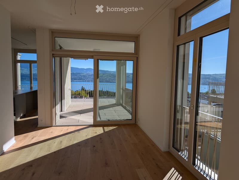 Bright, empty room with wooden flooring and large glass doors leading to a balcony with scenic views of a lake and mountains.