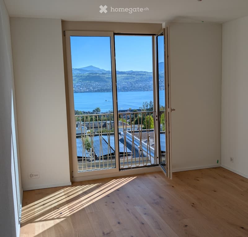 Empty room with wooden flooring, large sliding glass doors opening to a balcony with a scenic lake and mountain view.
