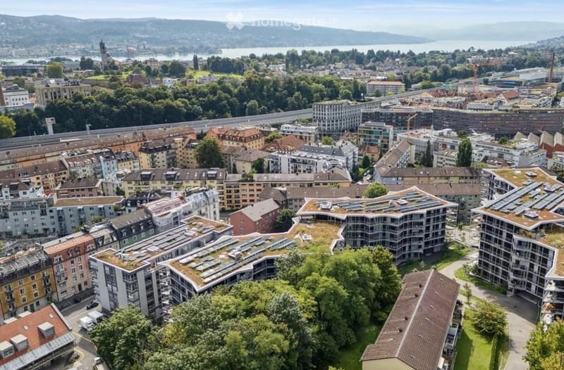 Aerial view of a cityscape with a mix of residential and commercial buildings, green spaces, and a body of water in the background.
