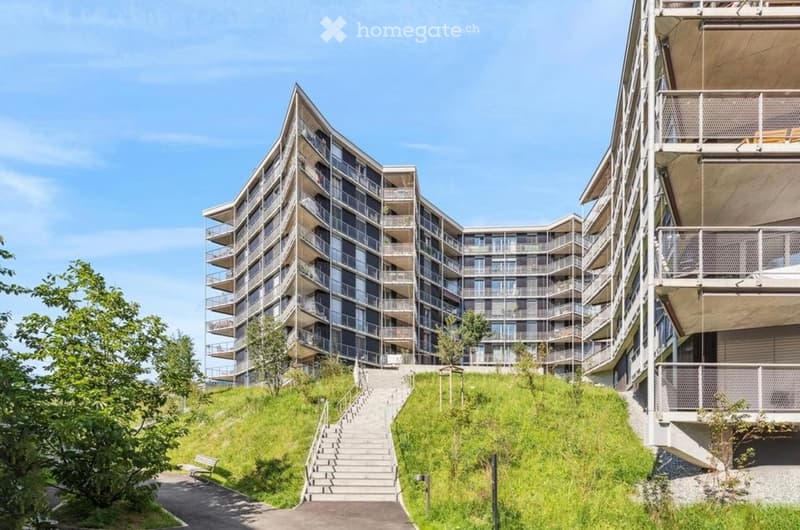 Modern multi-story residential building with balconies and large windows, situated on a raised ground floor with a staircase leading up to the entrance.