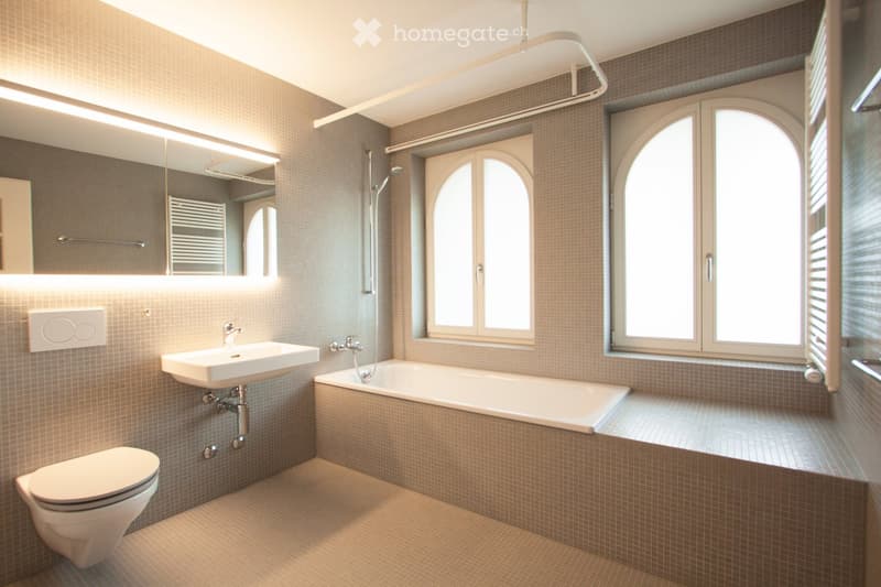 Modern bathroom with beige tiles, a white sink, bathtub, toilet, and large arched windows allowing natural light.