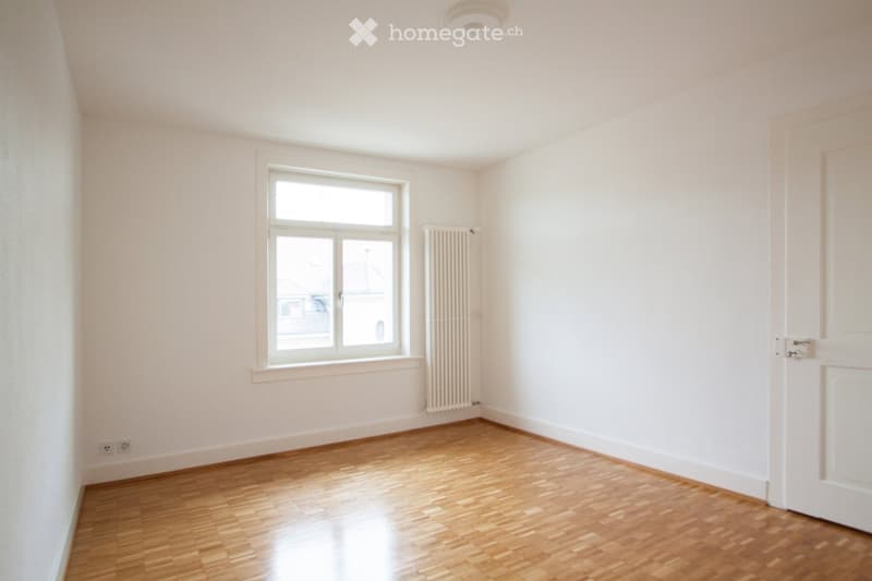 Empty room with white walls, wooden floor, single window, radiator, and door.