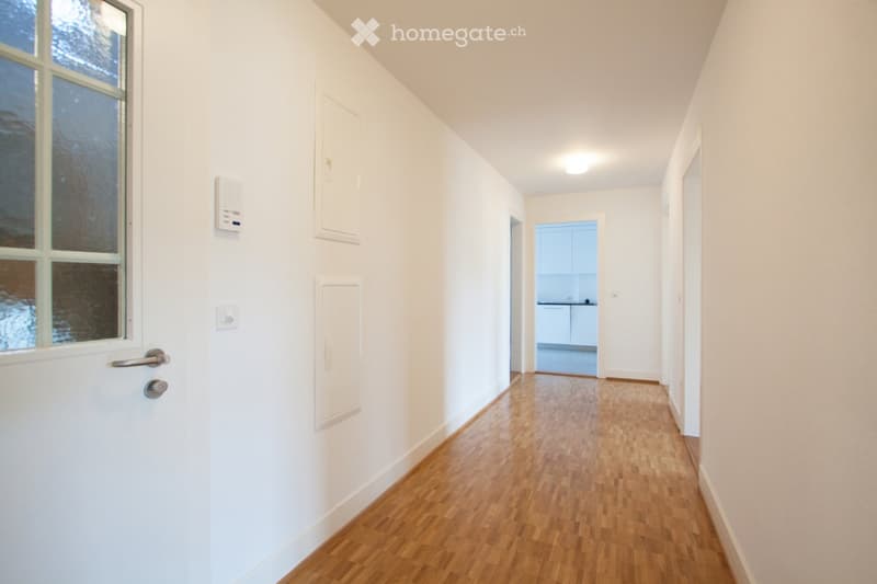 A clean, unfurnished hallway with white walls, wooden flooring, and a window on the left side. A door leads to a kitchen area visible at the end of the hallway.