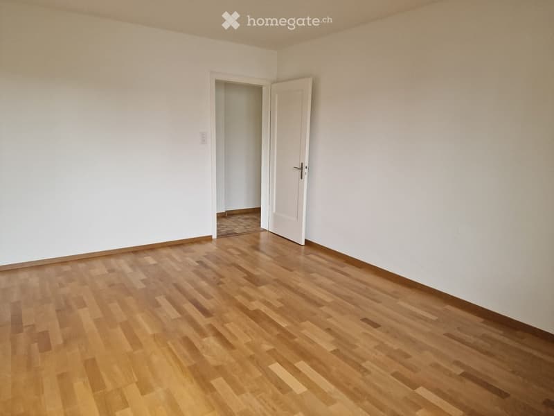 Empty room with light-colored wooden flooring and white walls, featuring an open doorway leading to another space.