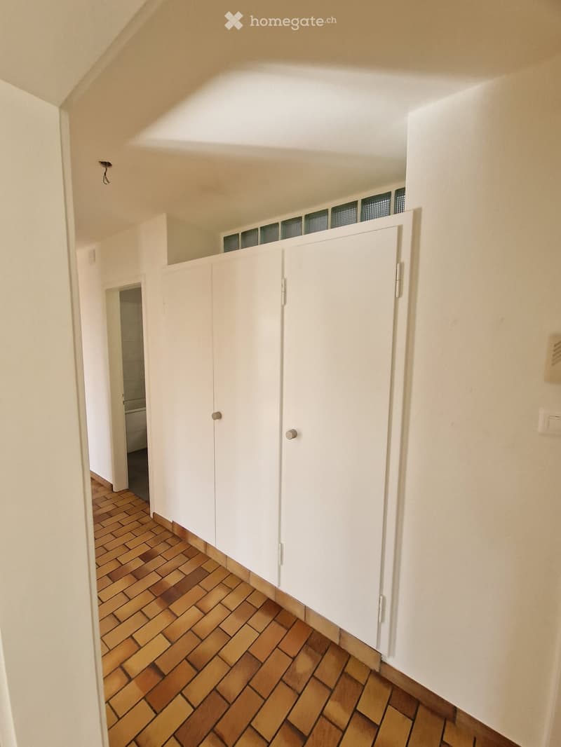A narrow hallway with white walls, wooden tiled flooring, and built-in white cabinets along one side. The hallway leads to another room with a visible doorway.