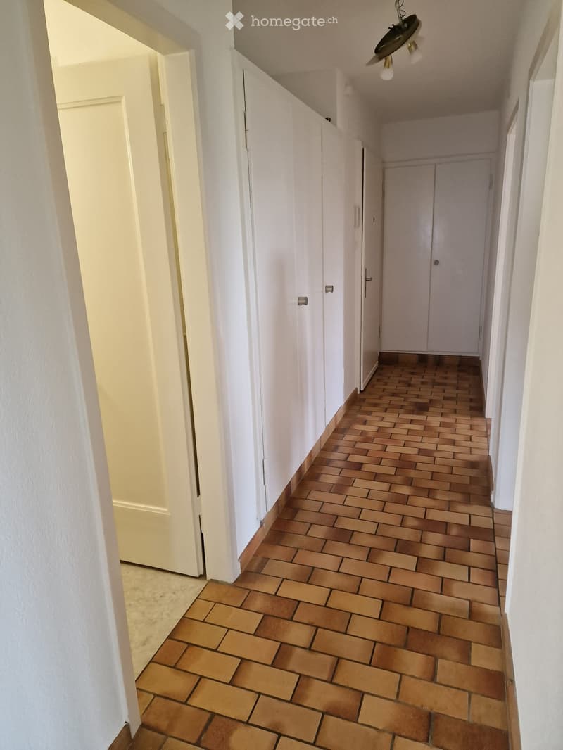 A long, narrow hallway with white walls and a patterned terracotta tile floor. Doors line both sides, and the ceiling features a simple light fixture.