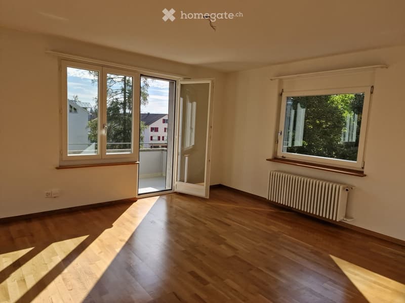 Empty room with wooden flooring, large windows, sliding glass door leading to a balcony, radiator, and wooden window sills.