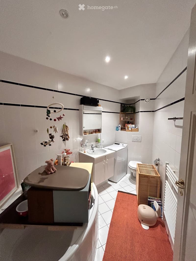 A compact bathroom with white tiled walls and floor, featuring a sink with a mirror cabinet, a toilet, a washing machine, a small wooden shelf, and a radiator. A toy and a stool are placed on the floor.