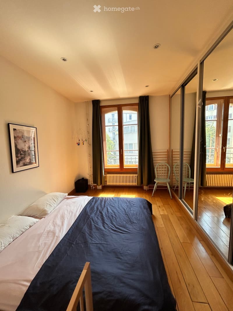 Cozy bedroom with wooden flooring, a single bed with blue bedding, two chairs, a picture frame on the wall, and a large window with curtains leading to a balcony.