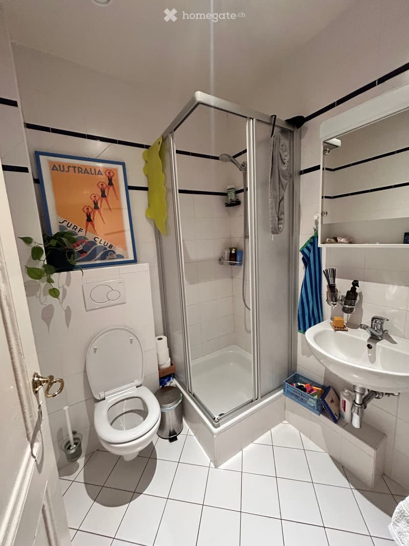 A modern bathroom with white tiles, a toilet, a sink, a shower, a mirror, and a framed poster on the wall.