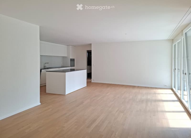 Empty room with light wooden flooring, white walls, and a small kitchen area with white cabinets and a countertop.