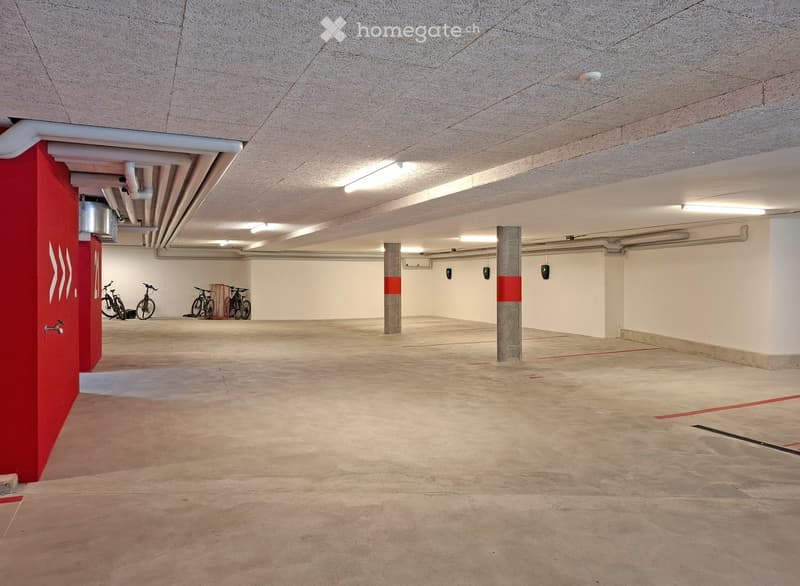 Underground parking garage with concrete floors, white walls, and several support pillars. Bicycles are parked in the corner, and the space is well-lit with fluorescent lighting.