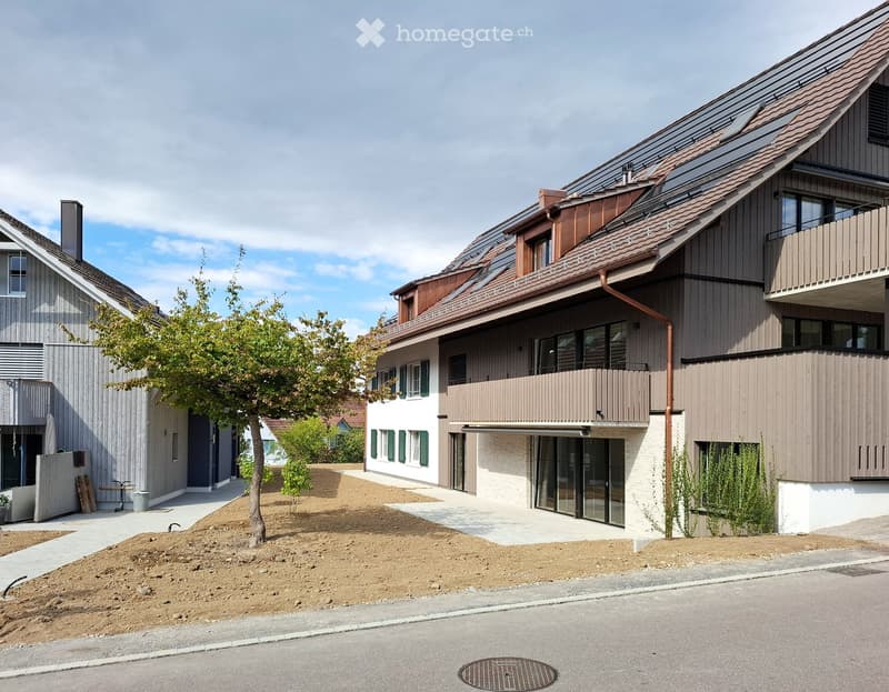 Two-story modern house with a sloped roof, large windows, and a balcony on the upper level. The house features a mix of wood and white siding, with a small garden area in front.