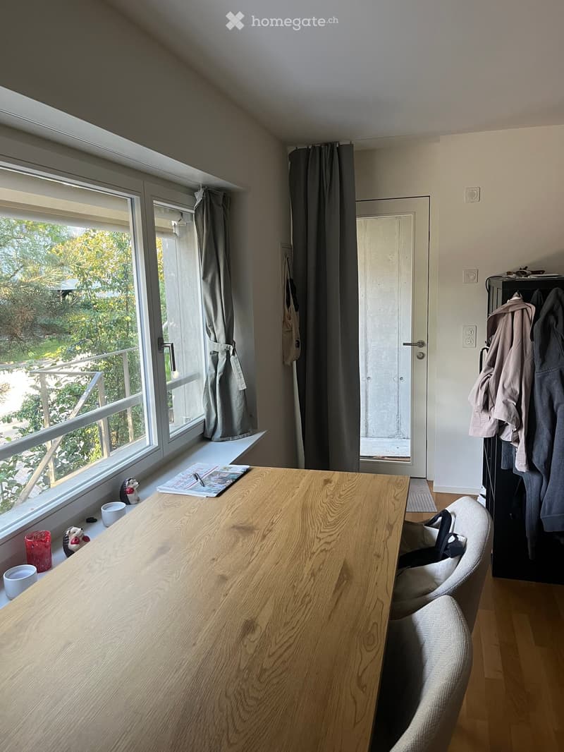 Bright room with wooden table, chairs, cups, toys, and a coat rack near the door. Large window with a view of greenery and a balcony.