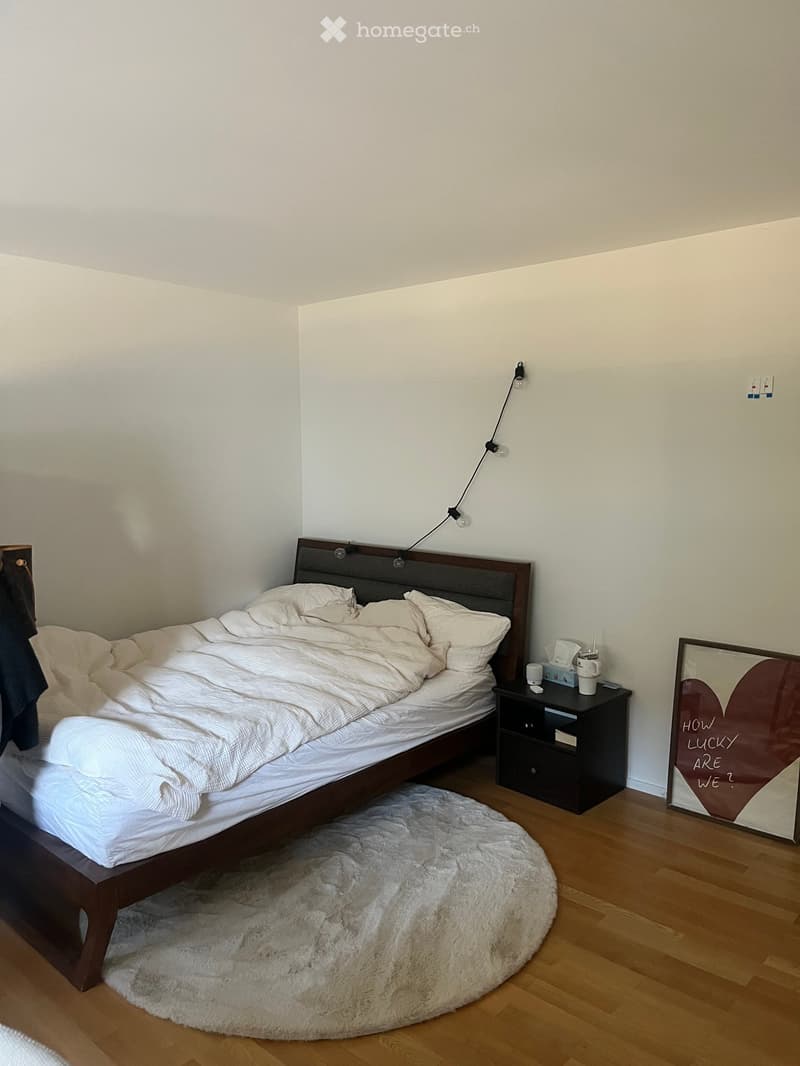 A modern bedroom with a wooden bed, white bedding, a nightstand with a lamp, a framed picture, and a fluffy white rug on the floor.