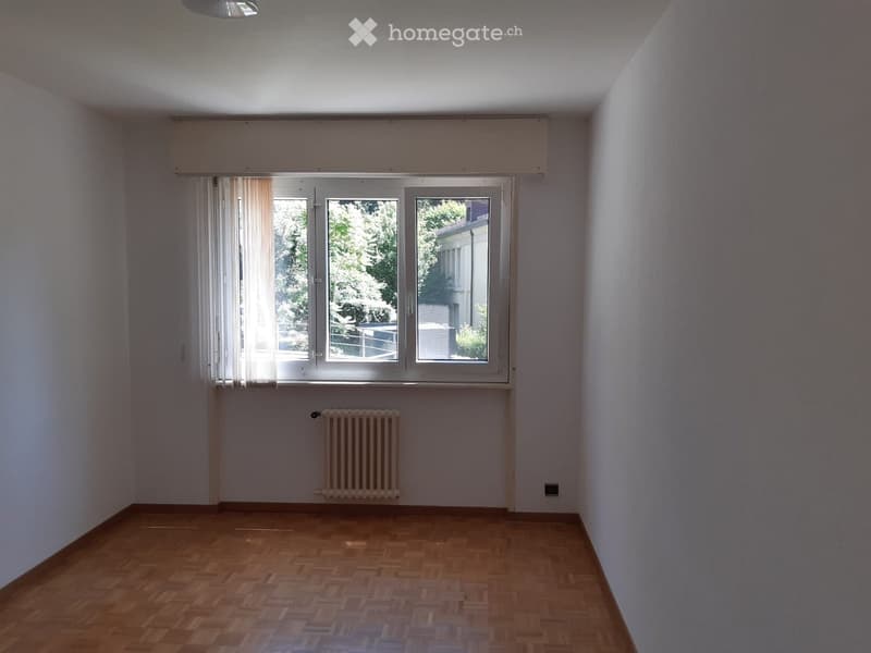 Empty room with wooden flooring, white walls, large window with blinds, and a radiator below the window.