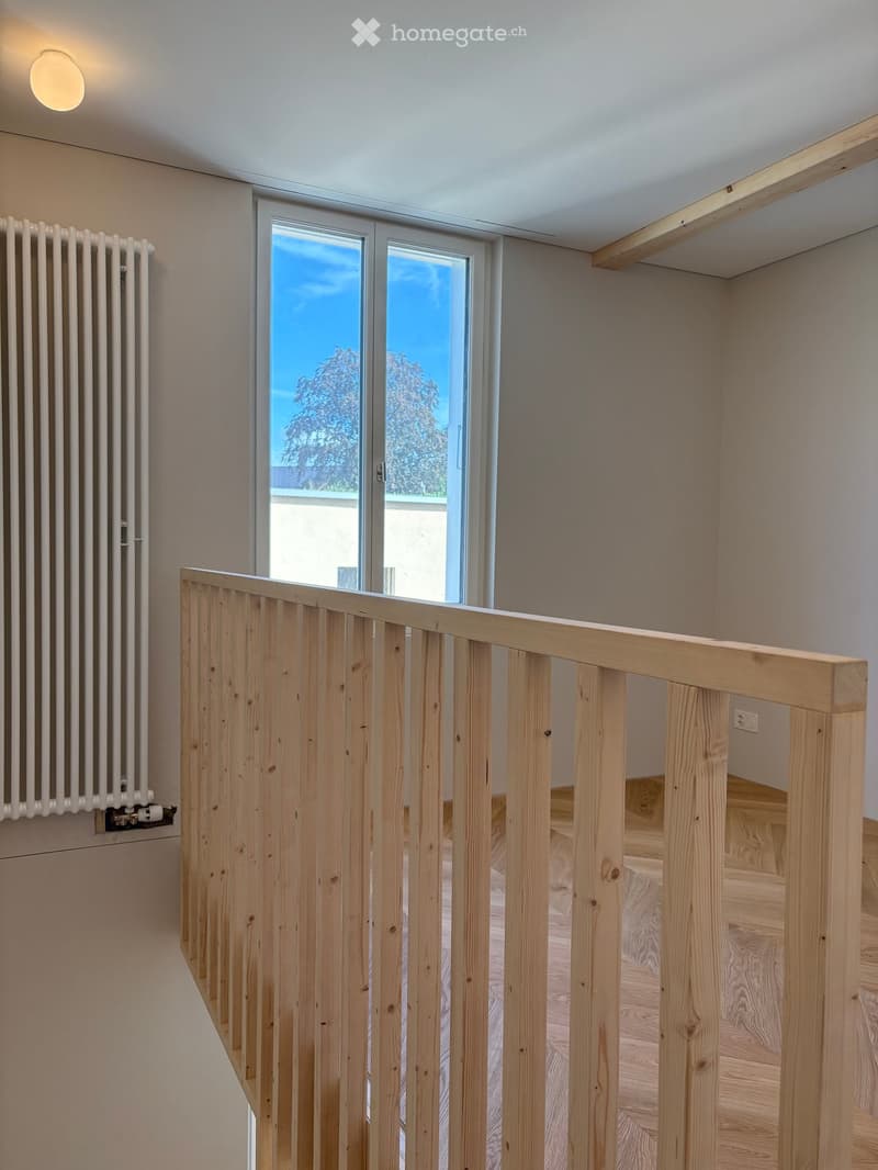 Modern hallway with wooden flooring, white walls, a wooden railing leading to an upper level, and a large window offering a view of trees and sky.