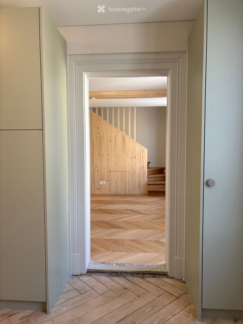 A modern hallway with herringbone-patterned wooden flooring, white walls, and a visible staircase in the background.