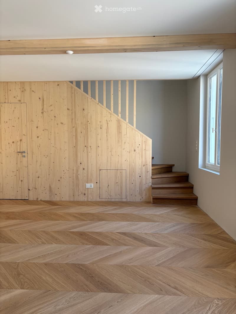 Modern hallway with wooden herringbone flooring, a staircase leading upwards, and a wooden door on the left side.