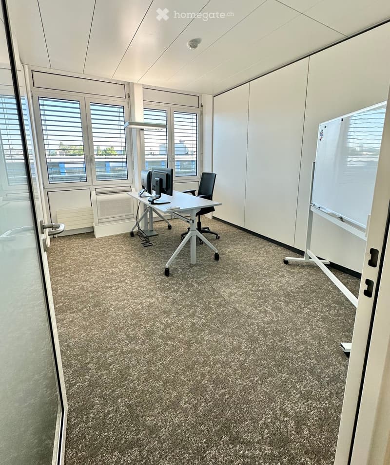 Office space with a desk, chair, computer, and whiteboard, large windows with blinds, and carpeted floor.