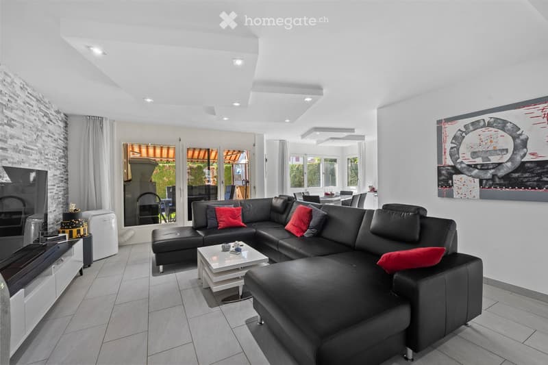 Modern living room with a black leather sectional sofa, red throw pillows, a white coffee table, a TV stand, and a fireplace. Large windows provide natural light, and there is an adjacent dining area visible through an open doorway.