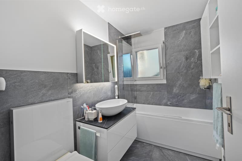 Modern bathroom with a white sink, bathtub, large mirror, and built-in storage. Features grey tiles, a window, and a glass shower enclosure.