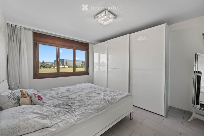 Modern bedroom with a white bed, pillows, a large wardrobe, and a mirror. The room features large windows with wooden frames and a view of the outdoors.