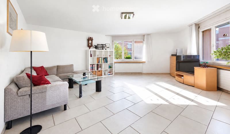 Bright living room with white walls, tiled floor, grey sofa with red cushions, glass coffee table, bookshelf, floor lamp, and TV on wooden stand.