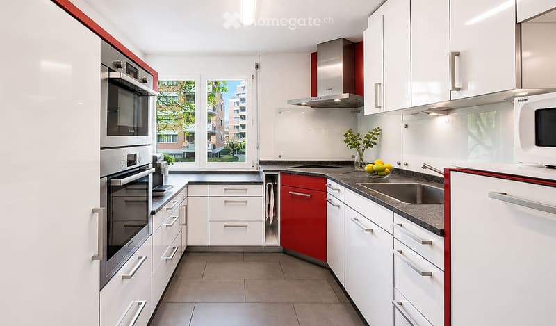 Modern kitchen with white cabinets, stainless steel appliances, and a red accent wall. Features include an oven, microwave, sink, and a bowl of fruit on the counter.