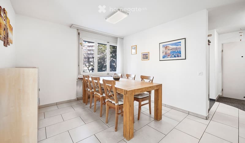 Bright dining room with a wooden dining table and chairs, large window with white curtains, and several picture frames on the walls.